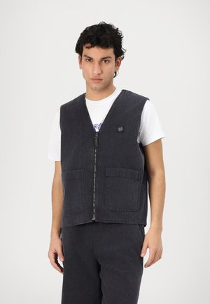 Dunkelgraue Cordweste mit V-Ausschnitt, Frontreißverschluss und zwei aufgesetzten Taschen. Über einem weißen T-Shirt gestylt. Texturiertes Gewebe und lässiges Design.