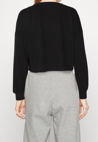 Pull court noir avec manches ballon longues, poignets côtelés et col rond, associé à un pantalon de survêtement gris clair avec détail de poche visible.