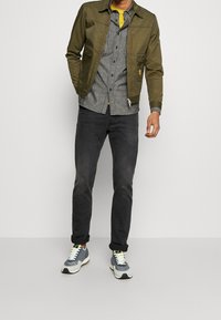 Uomo che indossa una giacca verde oliva sopra una camicia grigia a bottoni, jeans neri e sneakers grigie con suole bianche, in piedi contro uno sfondo neutro.