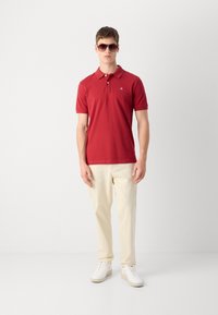 Rotes Polohemd mit Kragen und kurzen Ärmeln, kombiniert mit hellbeigen Hosen und weißen Sneakers, getragen von einem Modell mit Sonnenbrille.