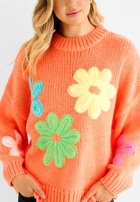 Bright orange knit sweater featuring large, textured flower appliqués in yellow, green, pink, and blue. Ribbed crew neckline and cuffs.