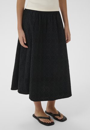 Vrouw draagt een gestructureerde zwarte midi-rok, een witte mouwloze top en zwarte teenslippers, staand op een effen achtergrond.