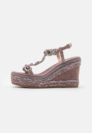 Sandalia cuña en ante rosa suave con tira adornada que presenta acentos metálicos y joyas; la plataforma trenzada añade textura y altura.