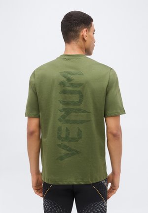 Mann trägt ein olivgrünes T-Shirt mit dunklem vertikalem "VENUM"-Schriftzug auf dem Rücken und schwarzen gemusterten Hosen, steht mit dem Rücken zur Kamera vor einem schlichten Hintergrund.