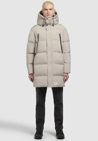 Beige puffertjakke med høj krave og justerbar hætte. Har to lynlåslommer og en let skinnende tekstur. Sorte accenter er inkluderet.