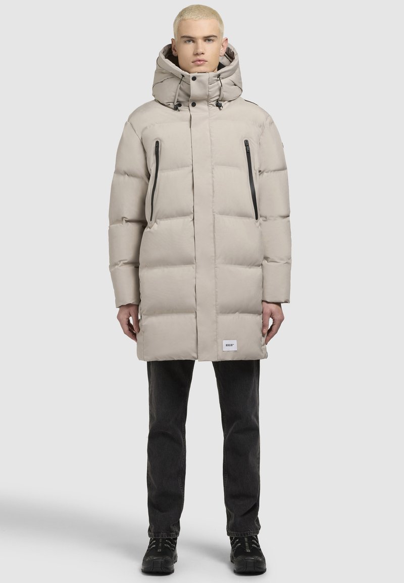 Beige puffertjakke med høj krave og justerbar hætte. Har to lynlåslommer og en let skinnende tekstur. Sorte accenter er inkluderet.