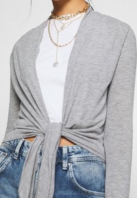 Grå lättviktswrap med knut fram, kombinerad med en vit topp och blå denimjeans. Accenterad med lager av guldfynd.