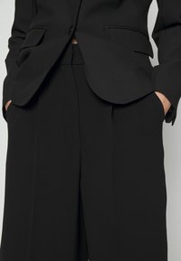 Person wearing a black tailored suit with hands in pockets, highlighting jacket and wide-leg trousers against a neutral background.