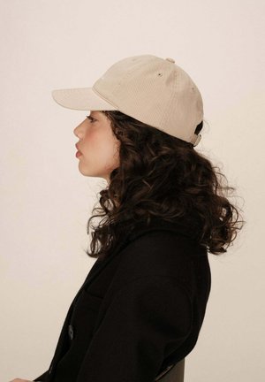Casquette de baseball en coton beige avec une visière incurvée, une sangle ajustable à l'arrière, et un tissu texturé. Le modèle porte un manteau noir.
