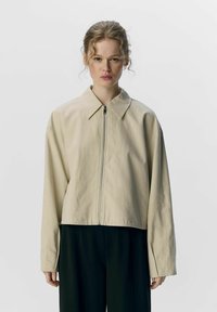 Giacca corta di un beige chiaro, realizzata in tessuto testurizzato, con colletto classico e zip frontale. Maniche lunghe e design minimale.