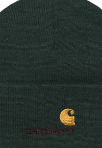 Zielona dziana czapka beanie z złotą haftowaną logotypem i czerwonym napisem. Tekstura jest prążkowana, co zapewnia dopasowane siedzenie.