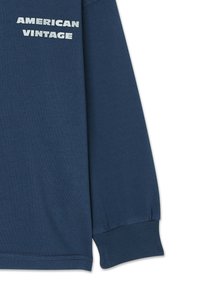 Navy blue long-sleeve shirt with ribbed cuffs. Features white print text "AMERICAN VINTAGE" on the front. Soft, cotton material.