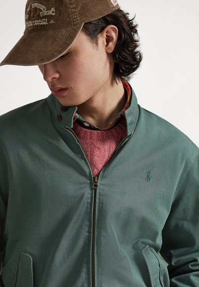 Πράσινο ελαφρύ μπουφάν με φερμουάρ μπροστά και τσέπες στα πλάγια, φορεμένο πάνω απ�ό ένα ροζ πλεκτό πουλόβερ και ένα πολύχρωμο πουκάμισο με γιακά.