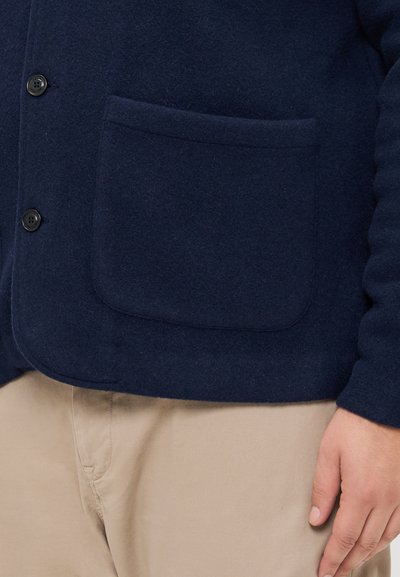 Δυοχρωμη μπλέ κάρντιγκαν από μάλλινο υλικό με μπροστινή τσέπη, διαθέτει κουμπιά και έχει απαλή υφή. Συνδυάζεται με μπεζ παντελόνι.