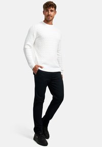 Hvit kabelstrikket genser med lange ermer, kombinert med mørke bukser og svarte joggesko. Genseren har et teksturert mønster.