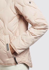 Padded pink jacket with quilted pattern, side pockets, and a zip closure. Features a black tag and a rounded collar.