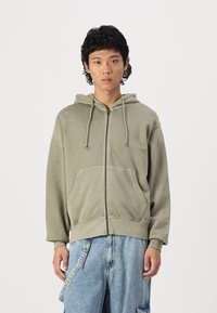 Olivgrüner Hoodie mit Reißverschluss, verstellbarem Kapuzenzug, zwei Fronttaschen und gerippten Bündchen. Der Stoff hat eine weiche, strukturierte Oberfläche.