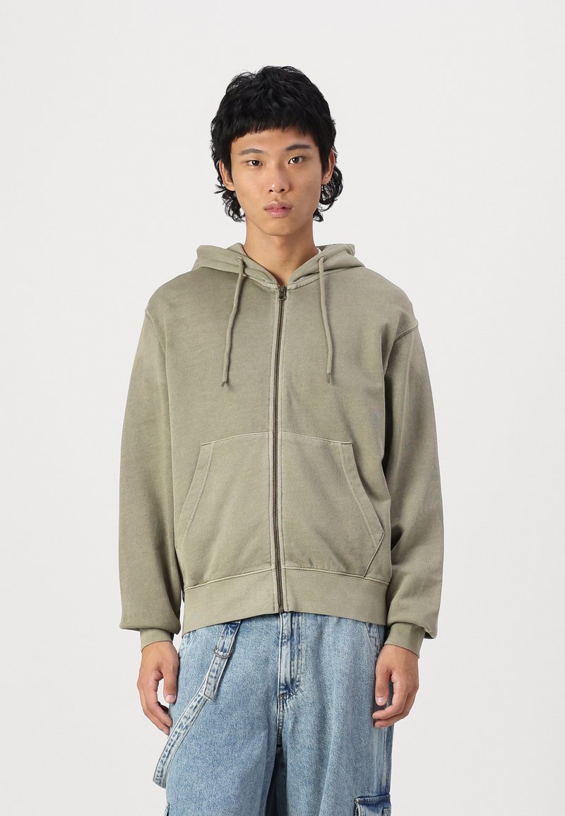 Olivgrüner Hoodie mit Reißverschluss, verstellbarem Kapuzenzug, zwei Fronttaschen und gerippten Bündchen. Der Stoff hat eine weiche, strukturierte Oberfläche.