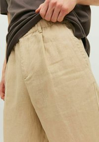 Pantaloni in lino beige chiaro con una vestibilità rilassata, dotati di tasche laterali e una texture sottile, abbinati a una maglietta marrone scuro.