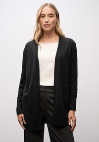 Svart, lång cardigan i mjukt tyg med ribbad textur och öppen framsida. Bärs över en vit topp och matchas med mörka byxor.