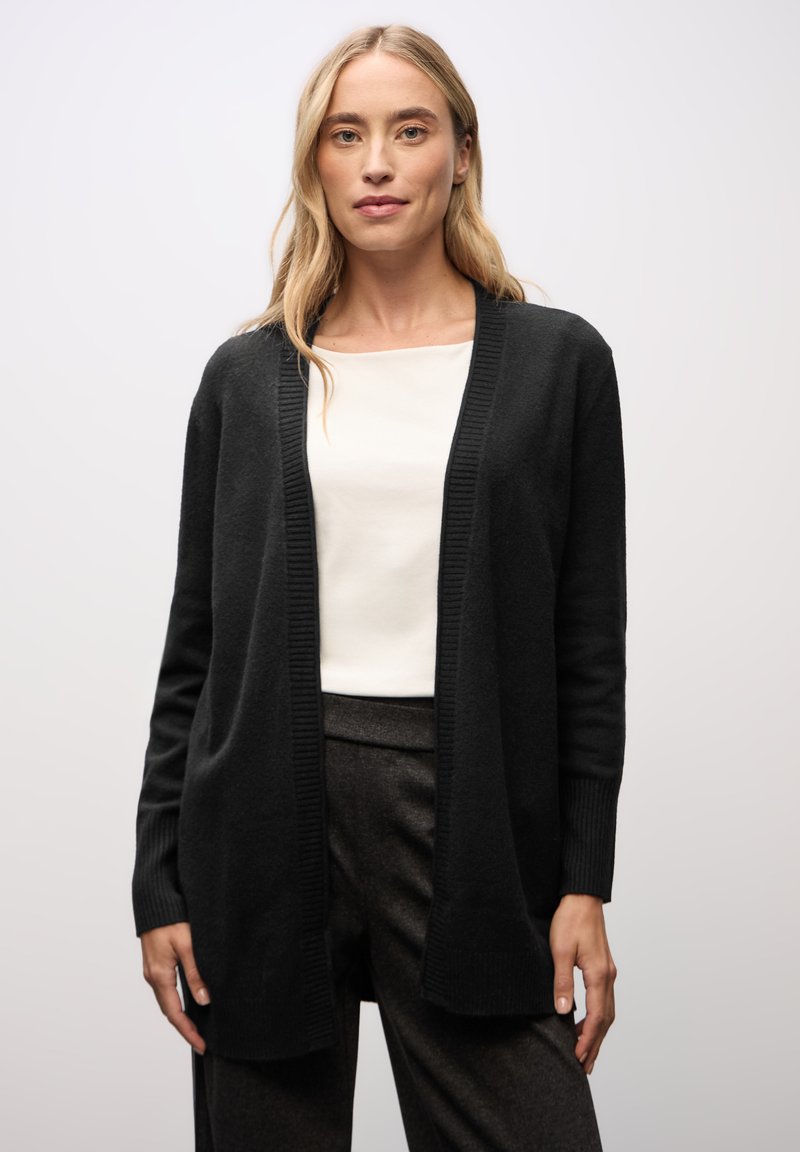 Svart, lång cardigan i mjukt tyg med ribbad textur och öppen framsida. Bärs över en vit topp och matchas med mörka byxor.