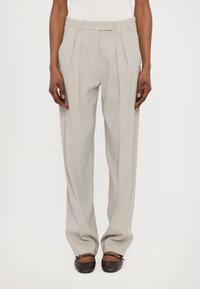 Lichtgrijze broek met een hoge taille, geplooide voorkant en een relaxte pasvorm. De stof lijkt glad met een subtiele textuur. De voeten zijn in donkerbruine schoenen.