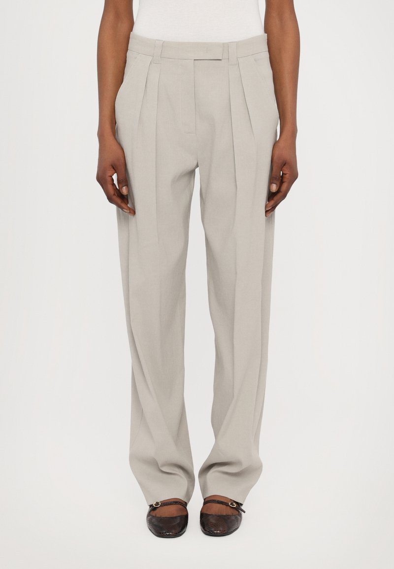 Lichtgrijze broek met een hoge taille, geplooide voorkant en een relaxte pasvorm. De stof lijkt glad met een subtiele textuur. De voeten zijn in donkerbruine schoenen.
