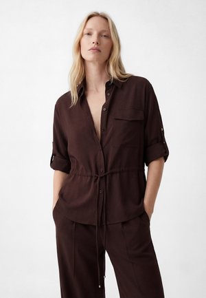 Femme blonde portant une chemise brun foncé à boutons avec manches retroussées et un pantalon assorti, debout les mains dans les poches sur fond blanc.