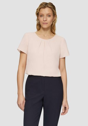 Blusa corta de manga corta en rosa claro con un escote redondeado y pliegues en la parte delantera, combinada con pantalones de un azul marino oscuro. Textura de tela suave.