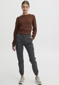 Brun teksturert genser med rund hals, kombinert med grå cargo-bukser med sidelommer. Modellen bruker lyse sneakers.
