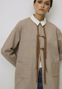 Beige visgraatjas met een witte kraag, voorstrikjes, twee zijzakken en een losse pasvorm. Getextureerde stof met een eigentijds ontwerp.
