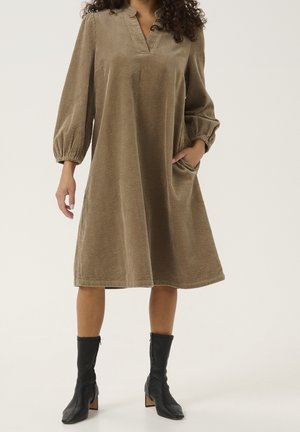 Femme portant une robe marron en velours côtelé, ample, longueur genou, manches longues et bottes noires à talons mi-mollet, main dans la poche.