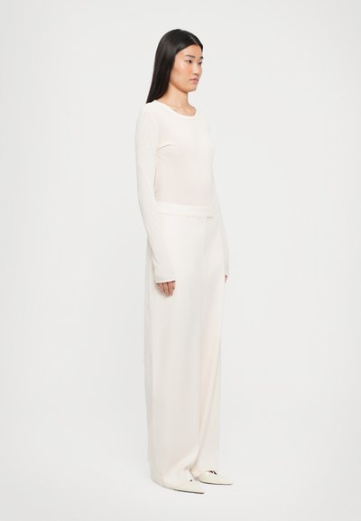 Donna con capelli lunghi e scuri, che indossa un completo tutto bianco, compresi una maglietta a maniche lunghe aderente, pantaloni larghi e scarpe con punta.