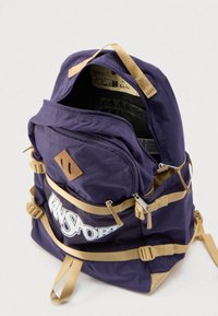 Σκούρος μπλε υφασμάτινος σάκος πλάτης με λουριά και λεπτομέρειες σε ταμπά χρώμα, διαθέτει μπροστινή θήκη με φερμουάρ, ρυθμιζόμενες αγκράφες στα πλευρά και λογότυπο στο κάτω μπροστινό μέρος.