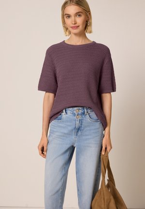 Kvinde iført lilla kortærmet striktop og lyseblå højttaljede jeans, der holder en brun tote-taske med den ene hånd ved siden.