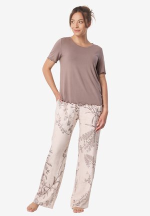 Kobieta stoi boso, ubrana w lawendową koszulkę z krótkim rękawem i luźne kremowe spodnie z botanicznymi nadrukami, na białym tle.