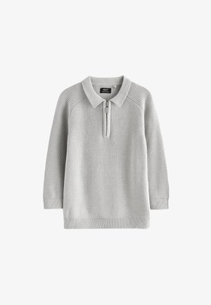 Mosolygós fiatal fiú zsebre tett kézzel, szürke cipzáras pulóvert, fekete nadrágot és fehér tornacipőt viselve, egyszínű háttér előtt áll.