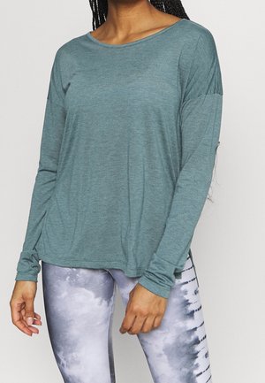Kobieta w luźnej turkusowej bluzce z długim rękawem i szarych legginsach tie-dye stoi na jednolitym tle.