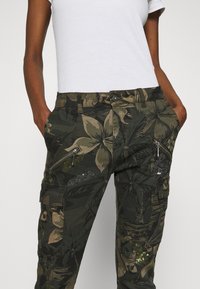 Pantalon cargo camouflage vert foncé avec motifs de feuilles, doté de poches zippées et d'accents en sequins. Confectionné en tissu robuste.