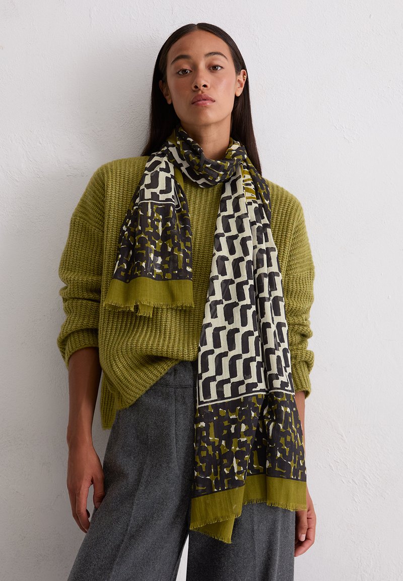 Pull vert en tricot porté avec une écharpe à motifs noirs et jaunes sur fond clair; l'écharpe a des bords frangés.