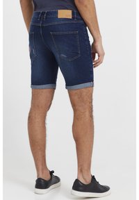 Traper šortsevi tamne boje, sa zarolanih rubovima, dva stražnja džepa i kožnom oznakom na pojasu. Uparen sa crnim tenisicama.