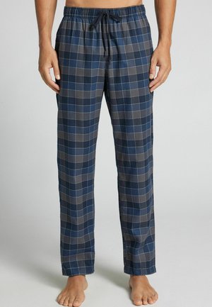Homme portant un pantalon de pyjama à carreaux bleu et gris avec une taille élastique et un cordon, debout pieds nus sur un fond uni.