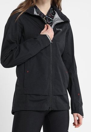 Person trägt eine schwarze Gore-Reißverschlussjacke über einem gemusterten Hemd und hat eine Hand am Reißverschluss in Brusthöhe.