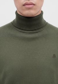 Gros plan d'une personne portant un pull à col roulé côtelé vert olive, montrant le cou et le bas du visage avec des lèvres neutres.