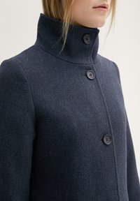 Mujer vestida con un abrigo de lana gris oscuro abotonado y un cuello alto, contra un fondo claro y simple.