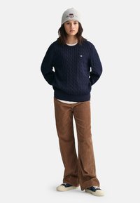 Marineblauer Kabelstrickpullover, braune Cordhosen, graue Mütze mit Logo, blaue Sneakers. Lässiges Outfit mit strukturierten Texturen und kontrastierenden Farben.