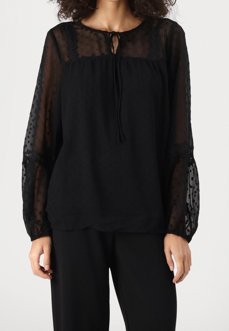 Femme portant un chemisier noir à manches longues avec un tissu transparent à pois sur les manches et le haut de la poitrine, col noué, associé à un pantalon noir.