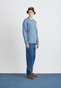 Jasnoniebieska koszula z długim rękawem z kieszonką, zestawiona z luźnymi niebieskimi dżinsami. Czarny kapelusz typu bucket i brązowe buty na sznurówki.