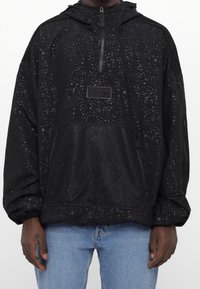 Svart huva tröja med spräckligt mönster och framficka i nät, bära med ljusblå jeans, visad från mittra bröstet till höfterna.