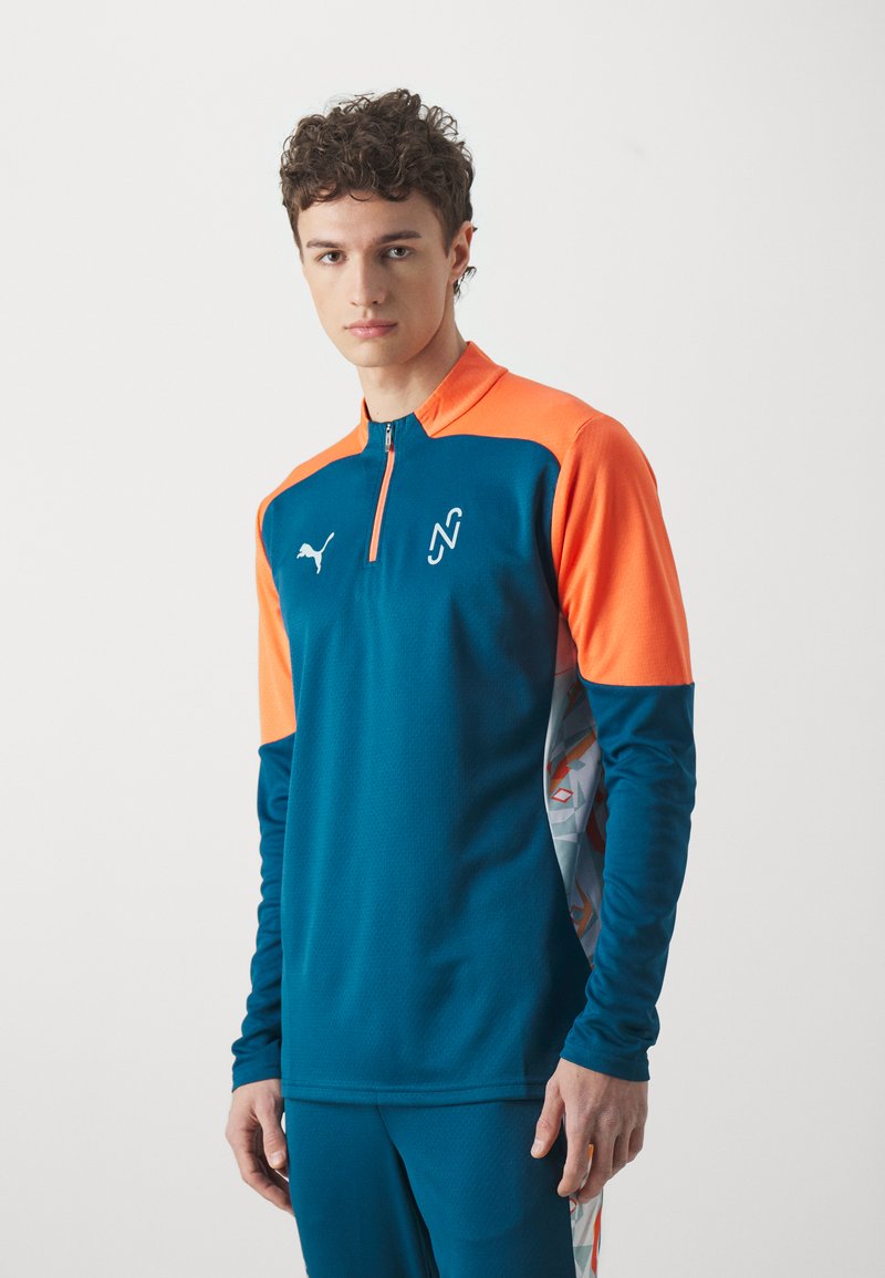 Top sportivo Puma in teal e arancione, con colletto a zip, maniche lunghe e pannello laterale con motivo geometrico. Tessuto morbido e vestibilità atletica.